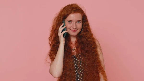 Redhead young woman in polkadot dress having pleasant mobile conversation using smartphone with friends, telling good news. Adult ginger freckles face girl indoors isolated on pink studio background