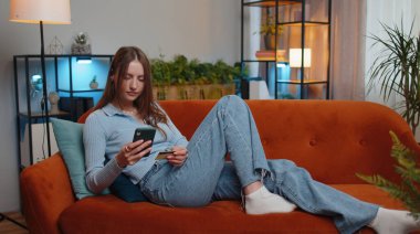 Genç bir kadın para transfer ederken kredi kartı ve akıllı telefon kullanıyor, online alışveriş yapıyor, modern bir evde yemek siparişi veriyor. Oturma odasında kanepede uzanan güzel kız.