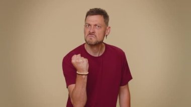 Aggressive angry handsome young man in red t-shirt trying to fight at camera, shaking fist, boxing with expression, punishment. Adult stylish male guy isolated alone on beige studio background indoors