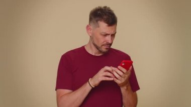 Sad young man in red t-shirt use smartphone typing browsing, loses becoming surprised sudden lottery results, bad fortune, loss. Adult stylish guy isolated on beige studio wall background indoors