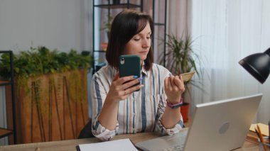 Kredi kartı ve akıllı telefon kullanan genç bir kadın para transfer ederken internetten alışveriş yapıyor. Ev ofisinin masasında oturuyor. Uzaktan kumanda işinde çalışan kız öğrenci.