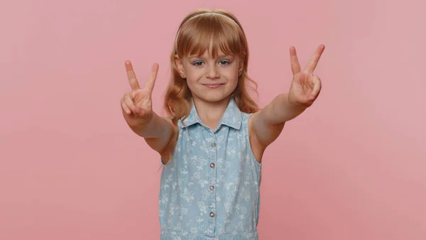 Happy preteen child girl kid showing victory sign, hoping for success and win, doing peace gesture smiling with kind optimistic expression. Little toddler children isolated on studio pink background