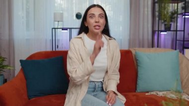 Shh be quiet please. Adult caucasian girl presses index finger to lips makes silence hush gesture sign do not tells gossip secret. Young woman at home apartment living room sitting on orange couch
