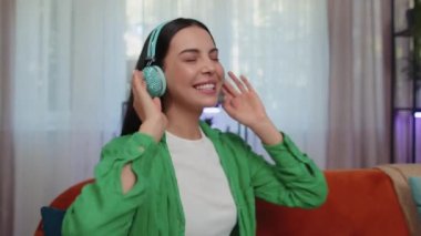 Neşeli, kablosuz kulaklıklı, evinde koltukta dans eden ve akıllı telefon eğlencelerinde en sevilen disko rock 'n roll müziğini dinleyen mutlu, gevşemiş genç kadın. İnsanlar hafta sonu aktiviteleri