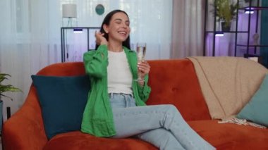 Joyful caucasian girl hold glass of champagne cheering drinking, celebrate success win, career grow up, lucky situation at home in living room. Young woman celebrating rent or purchase of new house