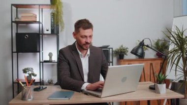 Businessman in suit working on laptop computer smiling friendly at camera and waving hands gesturing hello, hi, greeting or goodbye, welcoming with hospitable expression at office workplace desk