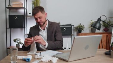 Sick ill businessman in suit suffering from cold flu or allergy working on laptop at home office. Manager freelancer man with towel on head blows sneezes wipes snot into napkin. Coronavirus quarantine