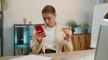 Businesswoman programmer software developer in suit working, making online purchase payment shopping with credit bank card and smartphone at modern office desk with laptop. Freelancer business woman