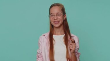 Smiling playful happy girl blinking eye, looking at camera with toothy smile, winking and flirting, expressing optimism. Young stylish female child kid isolated alone on blue studio background indoors