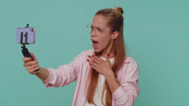 Wow reaction. Impressed pretty teenager girl student blogger taking selfie on mobile phone selfie stick, communicating video call online. Young stylish child kid on blue studio background indoor