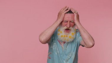 Senior man with flowers in beard screams from stress tension problems feels horror hopelessness fear surprise shock expresses gestures rage. Elderly grandfather isolated on pink studio wall background