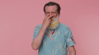 I am watching you. Senior man with flowers in gray-haired beard pointing at his eyes and camera, show I am watching you gesture, spying on someone. Elderly grandfather on pink studio wall background