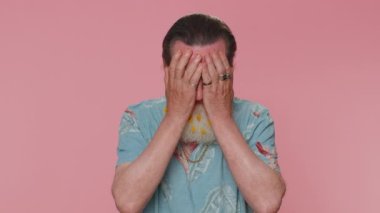 Upset disappointed senior man with flowers in gray-haired beard wipes tears and cries from despair, being sad because of unfair things. Elderly grandfather isolated alone on pink studio background