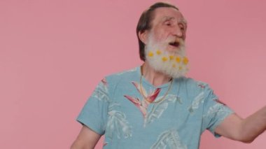 Smiling romantic senior man with flowers in beard sending lots of air kisses looking at camera and smiling, missing you, love confession. Elderly grandfather isolated alone on pink studio background