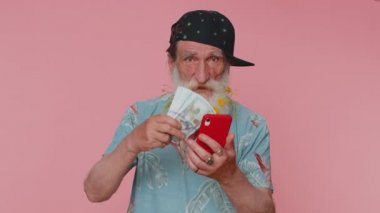Senior man with flowers in gray-haired beard looking smartphone display sincerely rejoicing win, receiving money dollar cash banknotes, success lottery luck. Elderly grandfather alone on pink wall