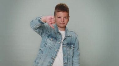 Upset unhappy teen boy in jacket showing thumbs down sign gesture, expressing discontent, disapproval, dissatisfied, dislike. Young children teenager child kid. Indoor studio shot on gray background