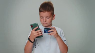 Sincere toddler boy customer using credit bank card and smartphone while transferring money, purchases online shopping, payment. Finance, internet. Young teenager child isolated on gray background