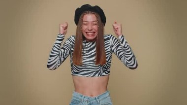 Happy pretty young woman shouting, raising fists in gesture I did it, celebrating success, winning, birthday, lottery goal achievemen. Adult stylish girl isolated on beige studio background indoors