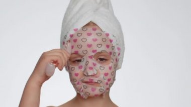 Pretty young child girl applying cosmetic moisturizing face mask, use massage roller tool. Teenager kid skin care treatment, natural cosmetics. Female close-up portrait on white background, indoors