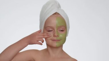 Beautiful young smiling child girl in towel on head applying cleansing moisturizing green facial mask. Teenager kid face skin care treatment, natural cosmetics. Female portrait. Perfect fresh clean