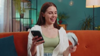 Planning family budget. Smiling happy girl counting money dollar cash, use smartphone calculate domestic bills at home. Joyful young woman satisfied of income saves money for planned vacation, gifts