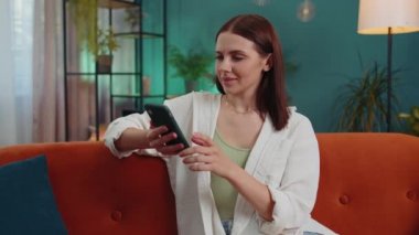 Portrait of adult girl sitting on sofa uses mobile phone smile at modern home apartment. Young woman texting share messages content on smartphone social media applications online, watching relax movie