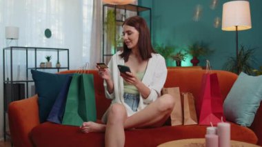 Happy shopaholic consumer girl sitting with shopping bags at modern home apartment making online order, payment with credit plastic bank card. Young woman on couch at home. Discount sale on internet