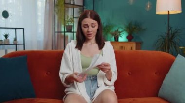 Stressed girl at home room looking at unpaid bank debt bills, doing paperwork, planning budget, calculate finances mortgage payments. Displeased young woman throws paper bills sitting on orange couch