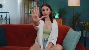 Stop gesture. Angry girl say No hold palm folded crossed hands, warning of finish, prohibited access, declining communication, body language, danger. Young woman at home room sitting on sofa indoors