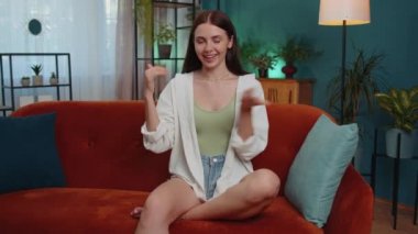 Portrait of caucasian cheerful rich girl showing wasting or throwing money around hand gesture, more tips earnings, big profit, win lottery, body language, share. Young woman at home in room on couch