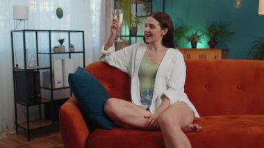 Joyful caucasian girl hold glass of champagne cheering drinking, celebrate success win, career grow up, lucky situation at home in living room. Young woman celebrating rent or purchase of new house