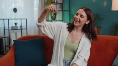 Young woman real estate agent lifting hand showing the keys of new home house apartment, buying or renting property, mortgage loan at modern home apartment indoors. Lovely girl in room sitting on sofa