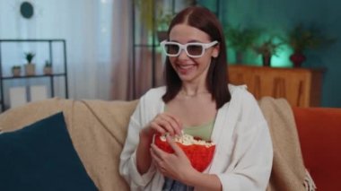 Excited adult girl sitting on sofa eating popcorn and watching interesting tv serial, sport game, film, online social media movie content online at home. Young woman enjoying domestic entertainment