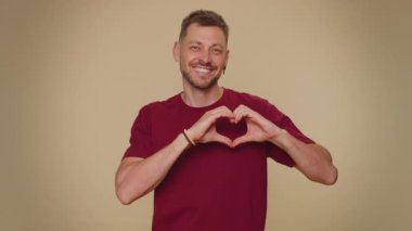 I love you. Smiling bearded handsome young man makes heart gesture demonstrates love sign expresses good feelings and sympathy. Adult stylish male guy isolated on beige studio background indoors