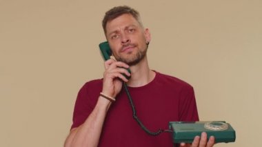 Crazy handsome young man in t-shirt talking on wired vintage telephone of 80s, fooling, making silly faces. Adult one stylish male guy isolated alone on beige studio background indoors. Call center