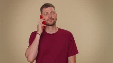 Tired young man in red t-shirt making online conversation speaks on smartphone. Boring call. Blah expression. Nonsense talking. Adult stylish male guy isolated alone on beige studio wall background