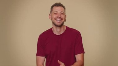 Portrait of happy bearded handsome young man in red t-shirt looking at camera, smiling. Success win. Adult stylish lovely male guy isolated alone on beige studio background indoors. Male nature beauty