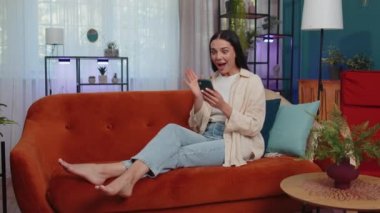Oh my God Wow. Excited happy joyful winner girl use smartphone typing browsing found out great win good news celebrate victory. Young woman with mobile phone lying at home in room on orange couch