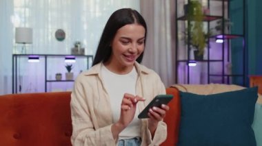 Portrait of lovely girl use mobile smartphone typing browsing say Wow yes found out great big win good news celebrate doing winner gesture. Young woman at home in living room sitting on orange sofa