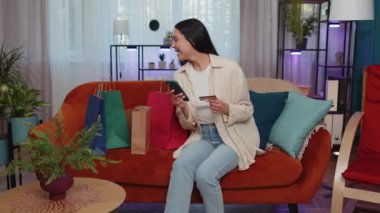 Happy shopaholic consumer girl sitting with shopping bags at modern home apartment making online order, payment with credit plastic bank card. Young woman on couch at home. Discount sale on internet