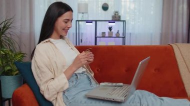 Girl using credit bank card and laptop computer while transferring money, purchases online shopping, order food delivery at home. Young woman sitting on couch holding dollar cash at home living room