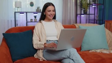 Portrait of blogger girl sitting on couch, looking at camera, making video webcam conference call with friends or family, enjoying pleasant conversation. Young woman laughing, waving hello at home