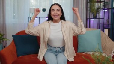 Happy caucasian girl shouting, celebrating success, winning, goal achievement, good news, raising fists in gesture I did it, lottery luck at home apartment. Young woman indoors in living room on couch