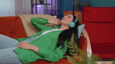Young woman in wireless headphones relaxing lying on sofa at home choosing listening favorite energetic disco rock n roll dancing music in smartphone. People weekend activities. Happy overjoyed girl