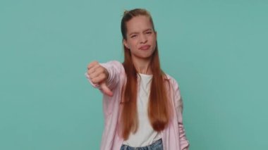 Upset lovely pretty teenager girl showing thumbs down sign gesture, expressing discontent, disapproval, dissatisfied, dislike. Young stylish female child kid isolated alone on blue studio background