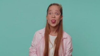 Cheerful funny bully teenager girl in shirt. Child girl showing tongue making faces at camera, fooling around, joking, aping with silly face, teasing isolated alone on blue studio background indoors