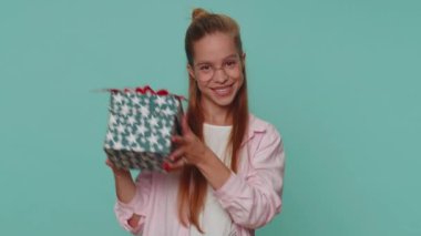 Positive smiling lovely pretty teenager girl presenting birthday gift box stretches out hands, offer wrapped present career bonus, celebrating party. Young child kid on blue studio background indoor