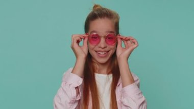 Portrait of seductive pretty teenager child kid wearing sunglasses, charming smile. Young stylish female teen student girl isolated alone on blue studio background indoors. People emotions concept