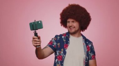 Tourist man in shirt traveler blogger taking selfie on mobile phone selfie stick communicating video call online with subscribers. Young adult guy boy 30s isolated alone on pink studio wall background