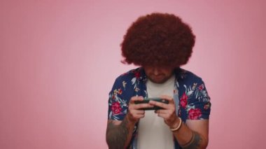 Endişeli, tişörtlü, komik bağımlı adam cep telefonuyla yarış oyunları oynuyor. Genç yetişkin adam pembe stüdyo arka planında izole edilmiş sürücü simülatörlü akıllı telefon aygıtı uygulaması kullanıyor